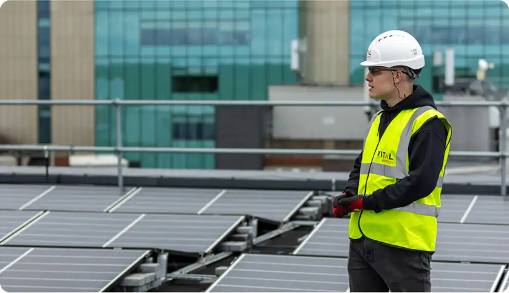 The image shows a young man, wearing a white hard hat, safety glasses, and a bright yellow safety vest with the logo "VITAL ENERGI" on it, standing on a rooftop. He is looking out at a large array of solar panels that cover most of the rooftop. In the background, a modern glass and concrete building is partially visible. The overall impression is one of renewable energy installation or maintenance.